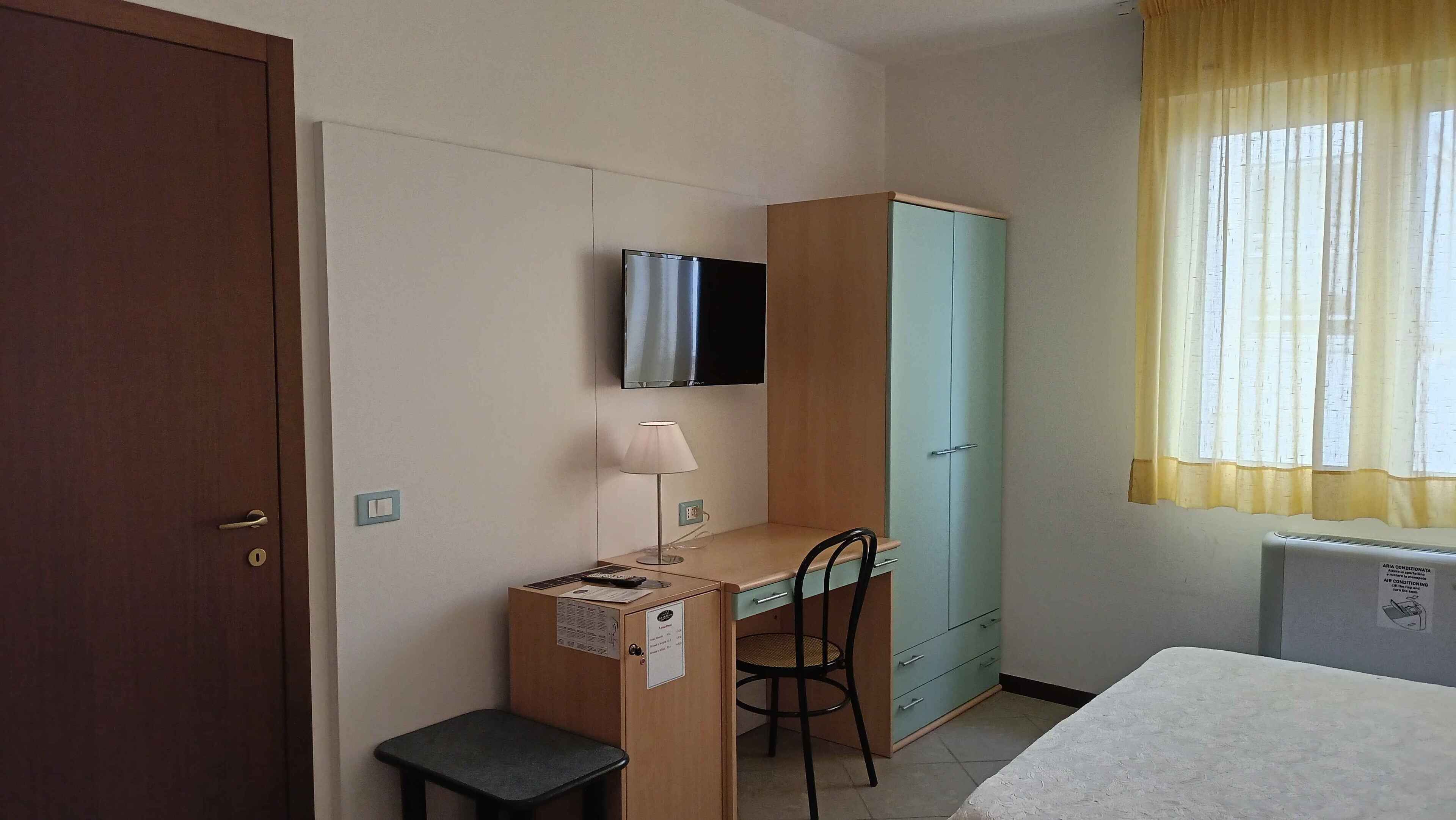 A small, simple room with a desk, chair, a television mounted on the wall, a cabinet, a small stool, and a window with curtains.