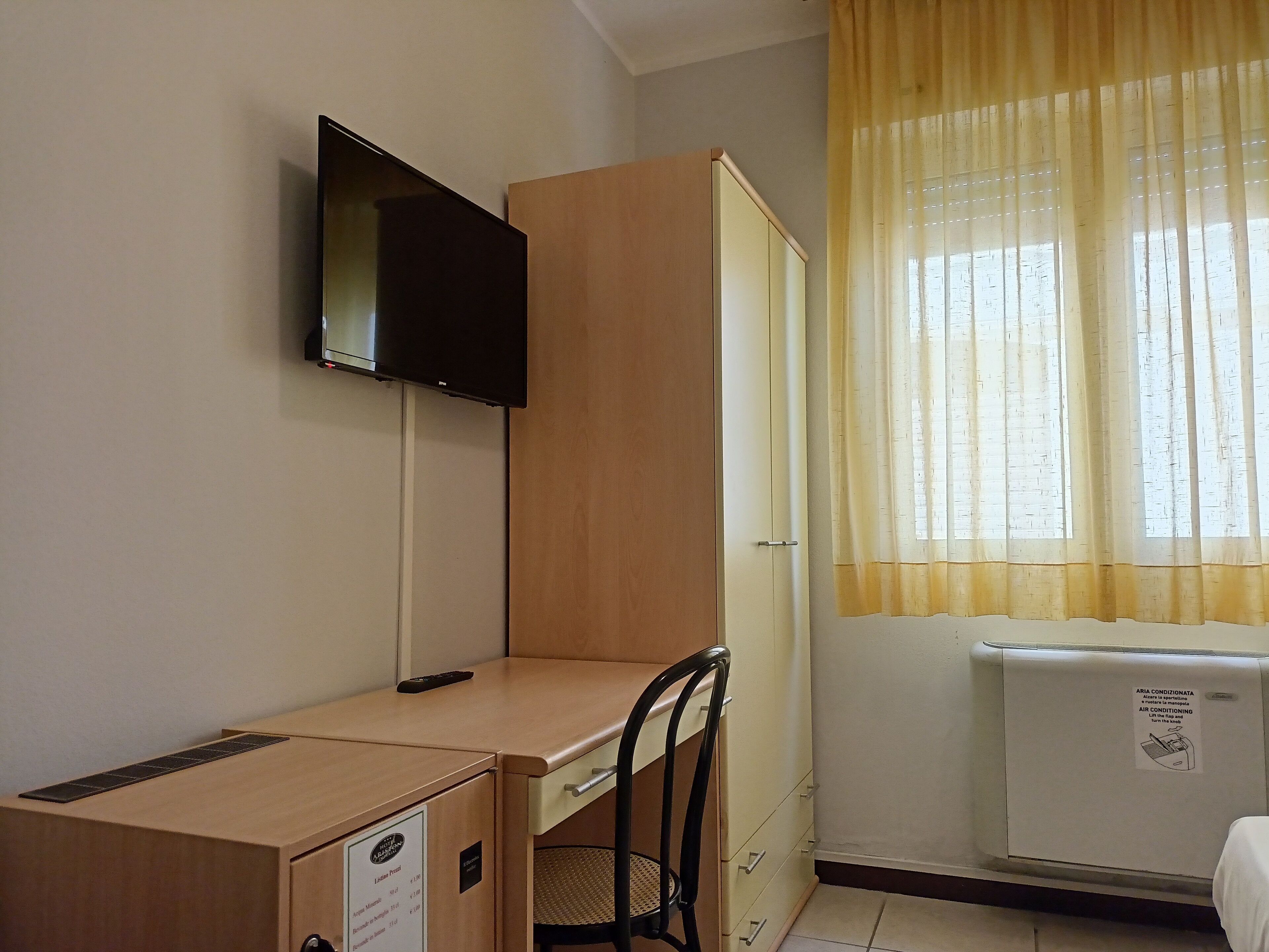 A small room with a desk, chair, a television mounted on the wall, and a window with curtains.