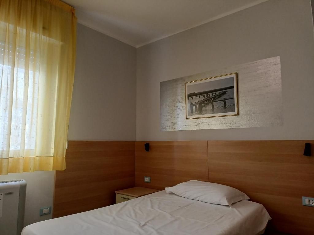 Camera d'albergo con un letto singolo, un comodino, un quadro appeso al muro e una finestra con tende.