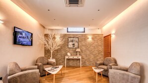 A modern waiting area with two sofas, a small table, a TV mounted on the wall, and a stone wall with a window. - Hotel Roma Tiburtina (Rome)