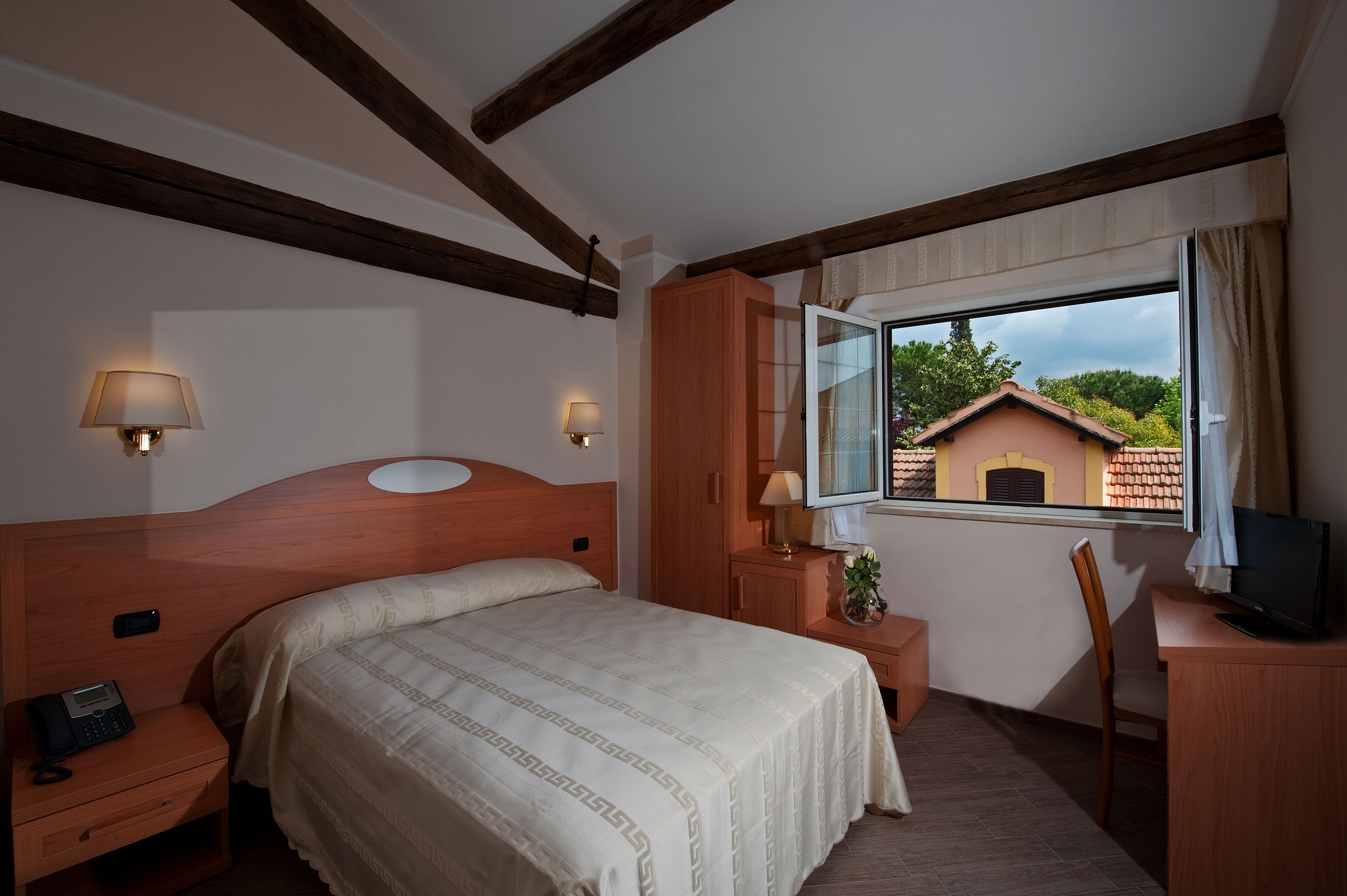 Ein Hotelzimmer mit einem Holzkopfteil, einem Bett mit einem gemusterten Überwurf, Nachttischlampen, einem Schreibtisch mit Fernseher und einem Fenster mit Blick auf ein Gebäude und Bäume.