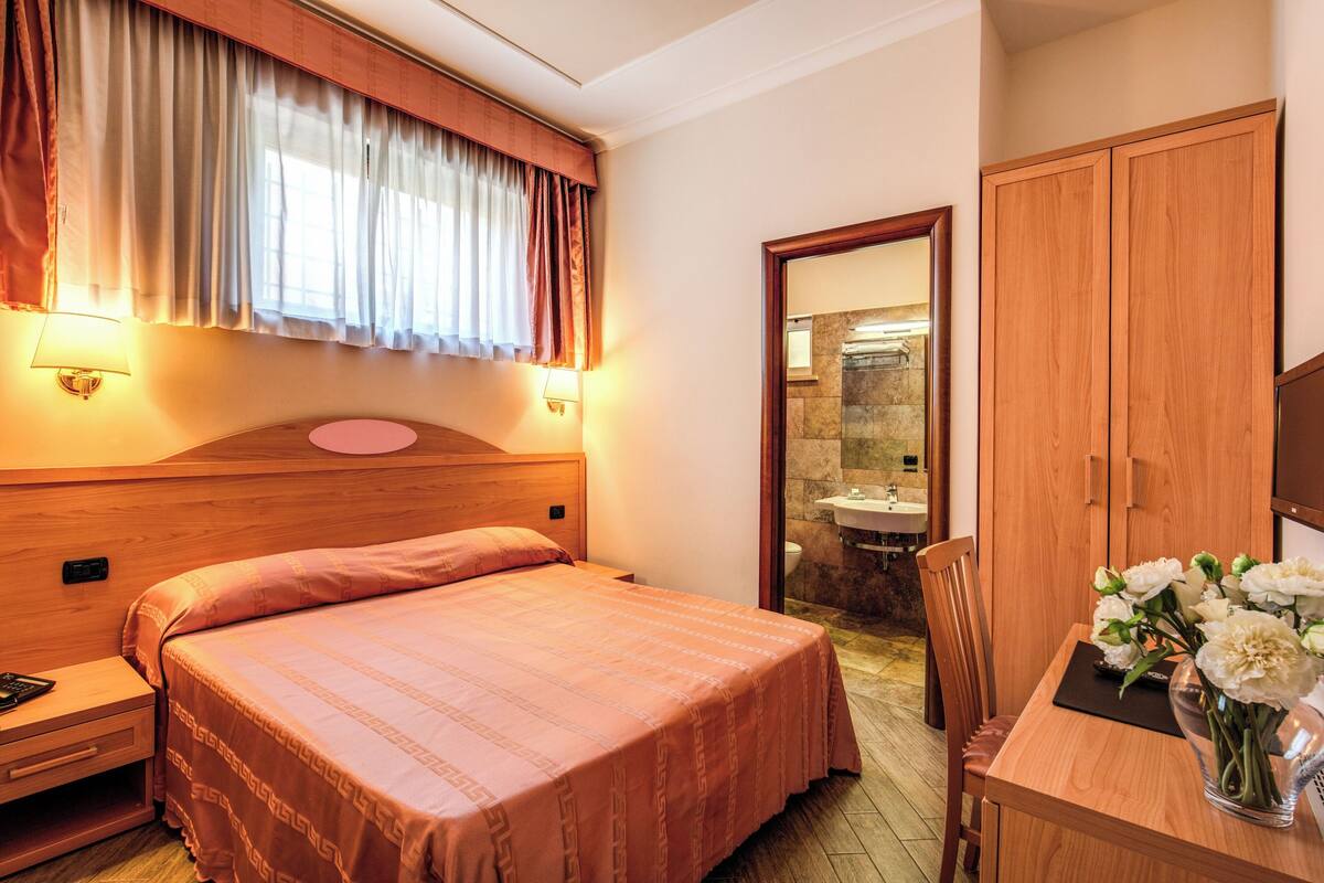 Ett hotellrum med en stor säng, ett träbeklätt sänggavel, ett skrivbord med en vas med blommor och ett badrum som syns genom en öppen dörr.