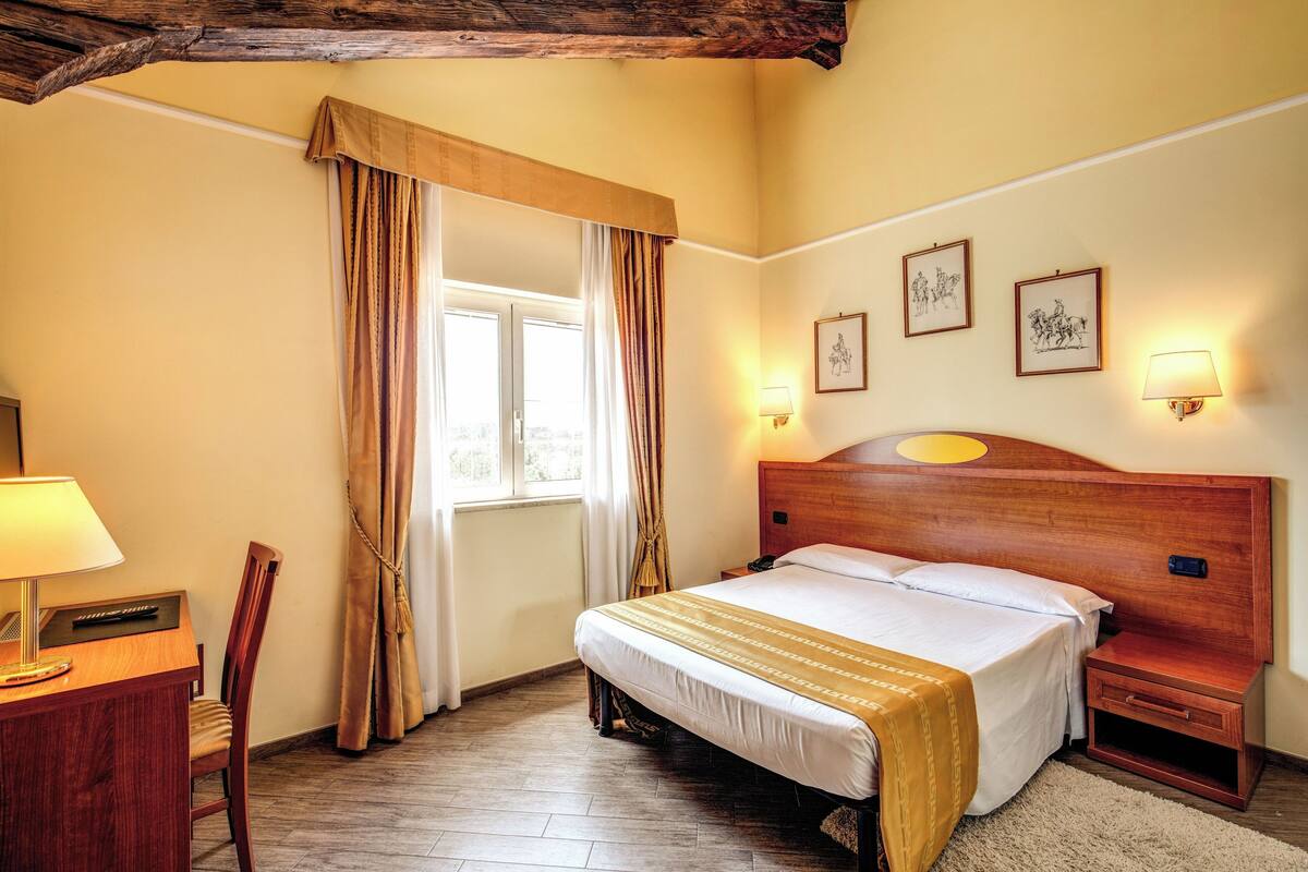 Ett hotellrum med en sänggavel i trä, en säng med ett vitt och guldskimrande överkast, ett skrivbord med en lampa, en stol och ett fönster med gardiner.