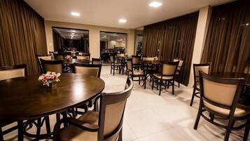 Uma ampla área de jantar com mesas e cadeiras redondas, um arranjo floral central e vista para a cozinha.