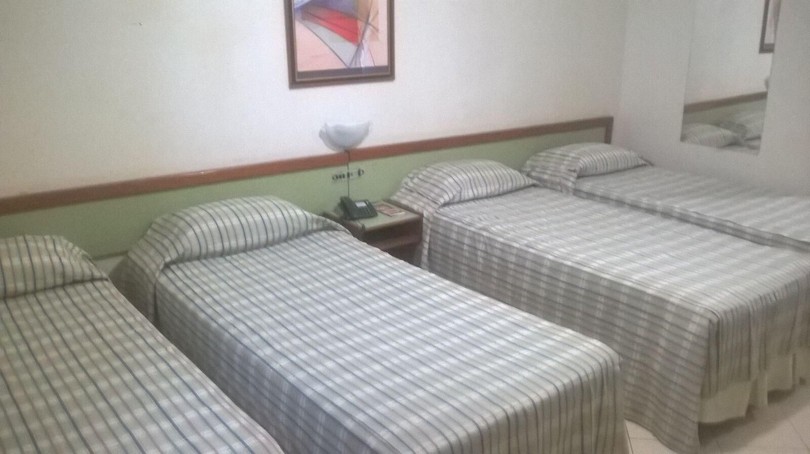 A hotel room with three single beds, a nightstand with a lamp, and a framed picture on the wall.