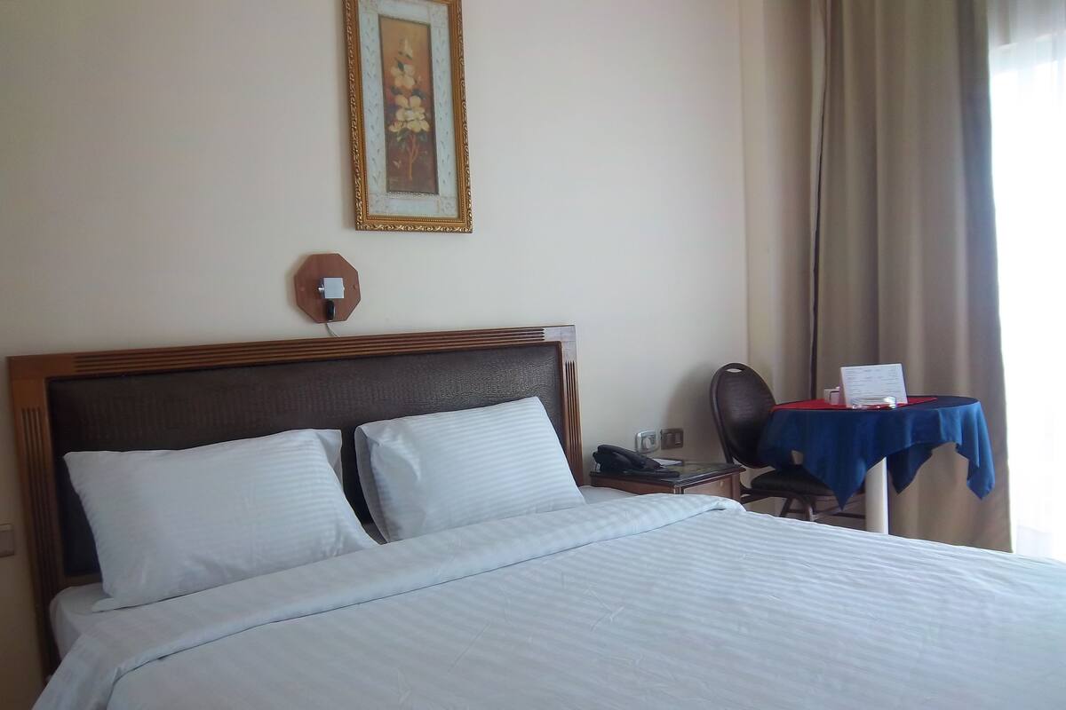 A hotel room with a large bed, a desk with a telephone and a menu, and a chair.