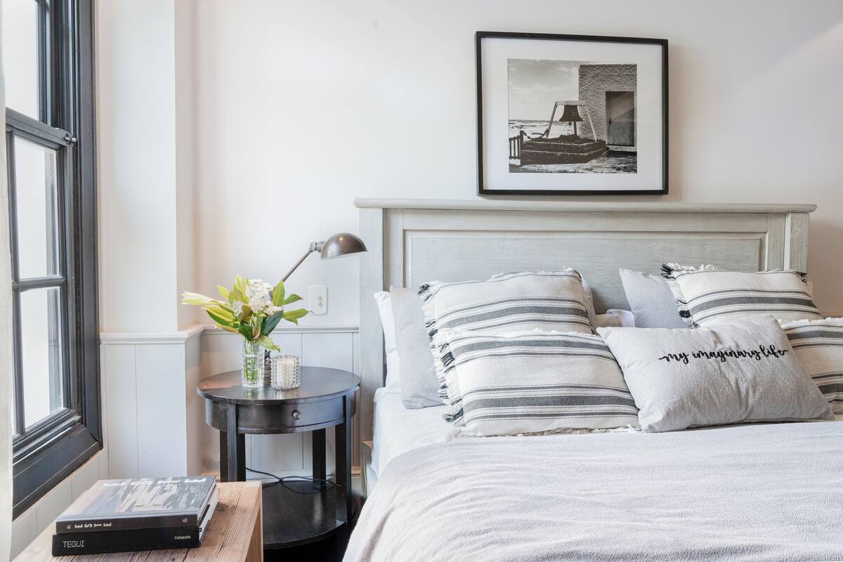 Une chambre bien rangée, avec un lit, des oreillers, une table de chevet avec une lampe, un tableau encadré au mur et une fenêtre.