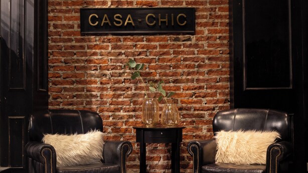 Une pièce avec un mur en briques, deux fauteuils en cuir, une petite table sur laquelle est posé un vase, et un sol à motifs.