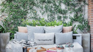 Un espace de détente extérieur accueillant, avec un banc rembourré, des coussins, une table avec des livres, une bougie et une plante en pot.