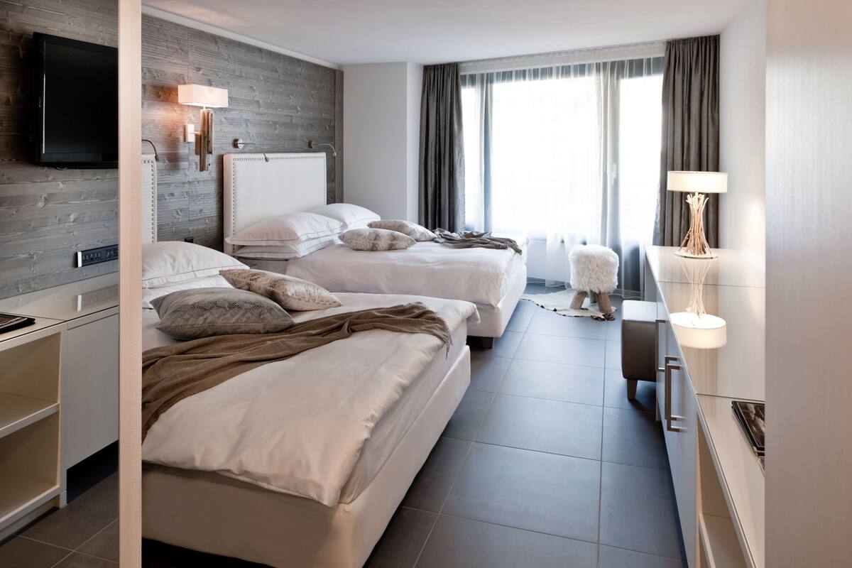 A modern hotel room with two beds, a flat-screen TV, a bedside table, a lamp, and a large window with curtains.