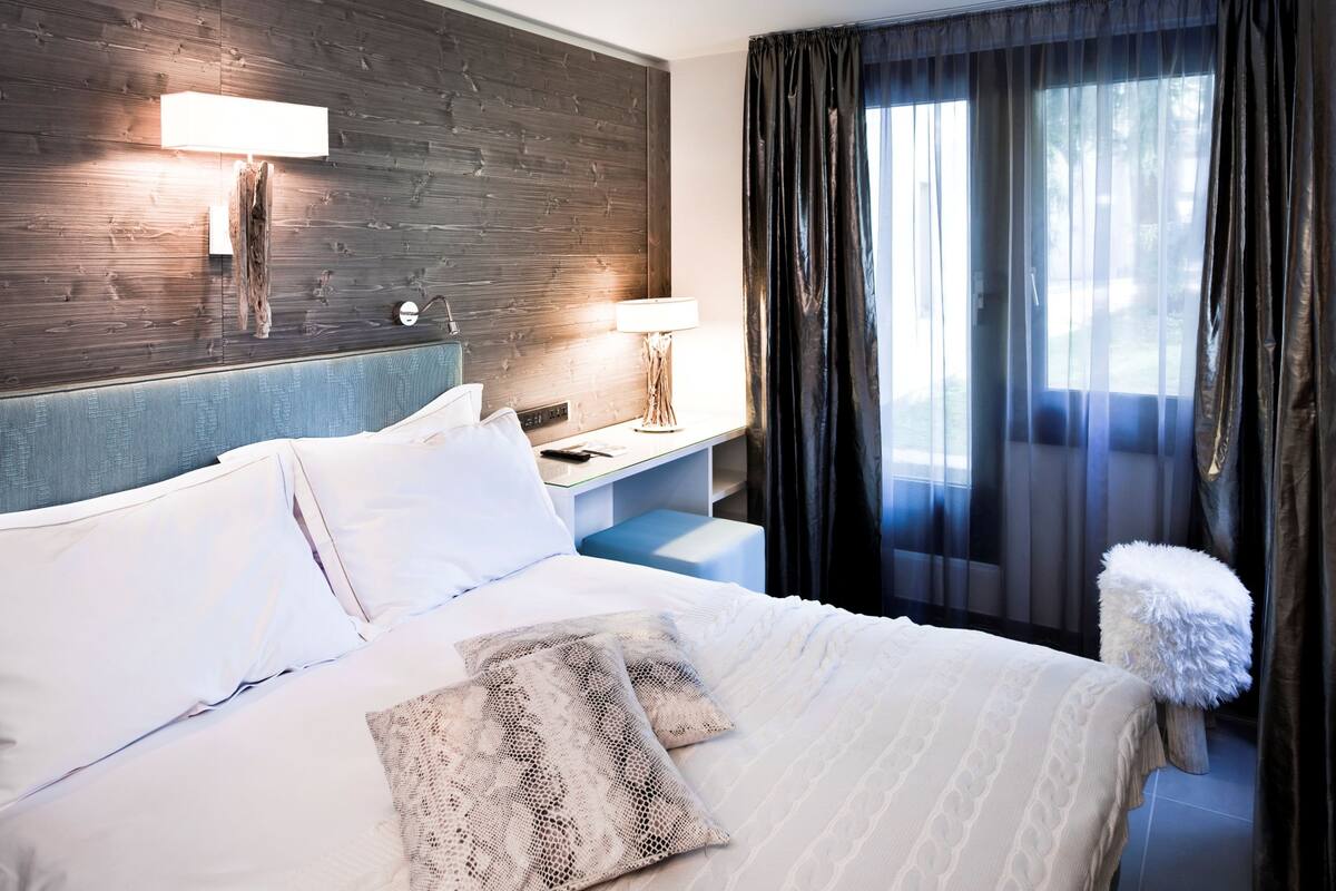 A hotel room with a large bed, a bedside table with a lamp, a wooden headboard, and a view of the outdoors through sheer curtains.