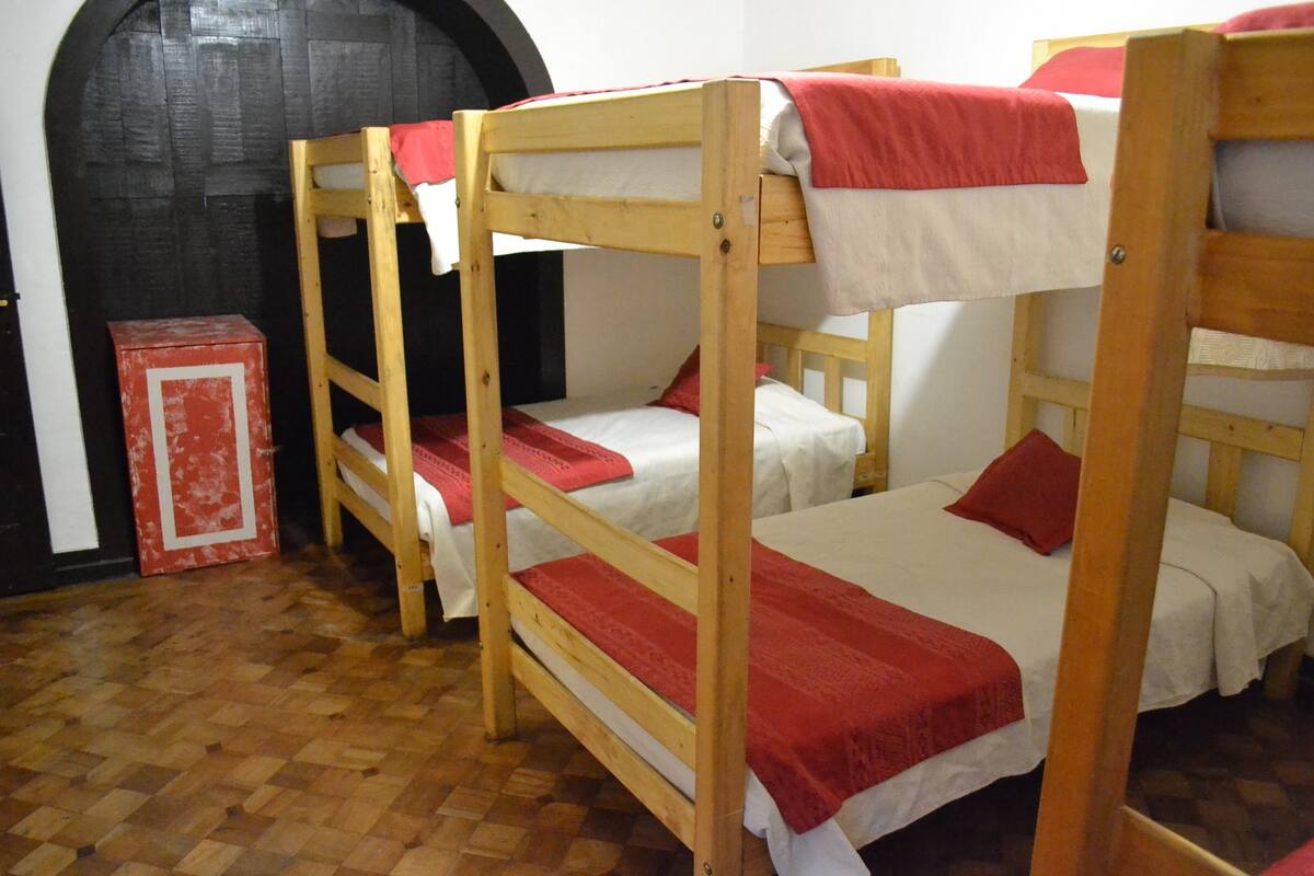 Una habitación con tres literas, cada una con una manta roja. Se observa un aparador de madera y una puerta negra al fondo.