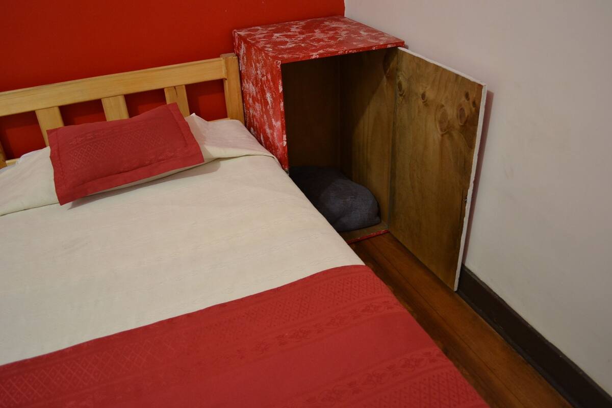 Una cama individual con una colcha roja y blanca, un almohadón rojo y un pequeño banco de madera con un paño rojo.