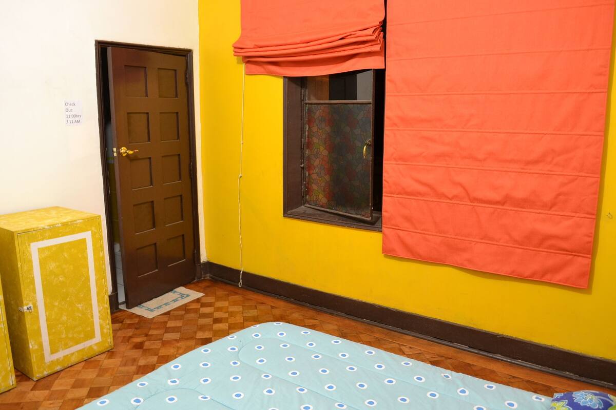 Una habitación pequeña con un letrero de recepción, una puerta, una ventana con cortina y una cama con colcha azul, junto a un mueble amarillo.