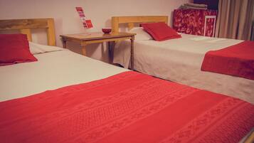 Habitación de hotel con dos camas, cada una con una manta y almohadas rojas, un cabecero de madera, una mesita con un bol y un libro rojo en la pared.