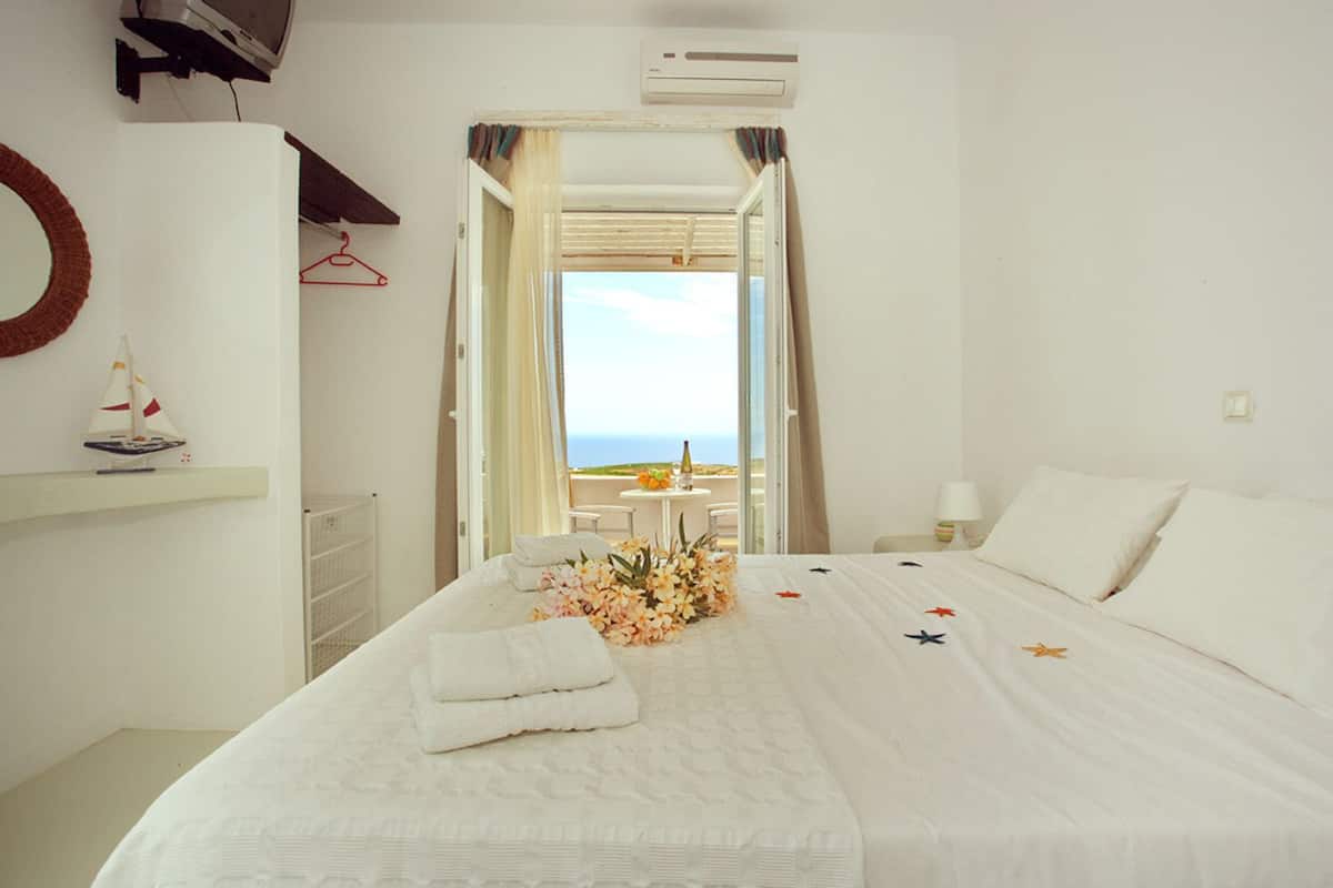 Ein Hotelzimmer mit Bett, einem kleinen Tisch mit einer Flasche, einem Stuhl und Blick auf das Meer durch eine offene Tür.