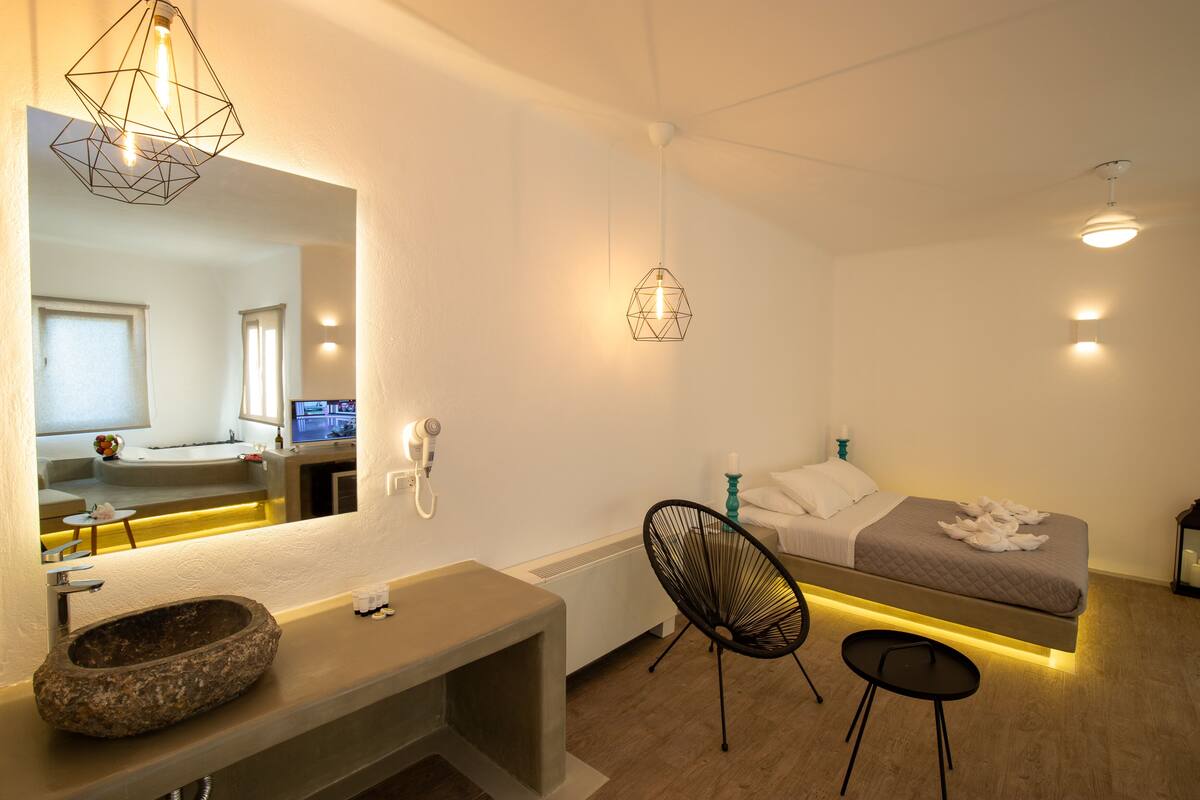 Ein modernes Hotelzimmer mit einem großen Spiegel, einem Becken aus Stein, einem Bett mit weißer Tagesdecke und einer geometrischen Pendelleuchte.