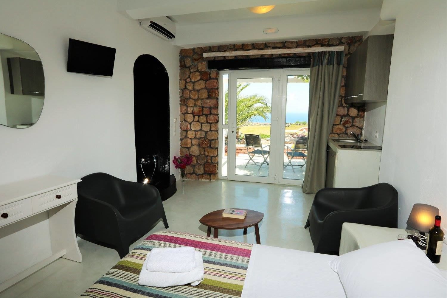 A hotel room with a bed, two chairs, a desk, a TV, a stone wall, a glass door leading to a balcony with a view, and a small table with a book and a bottle.