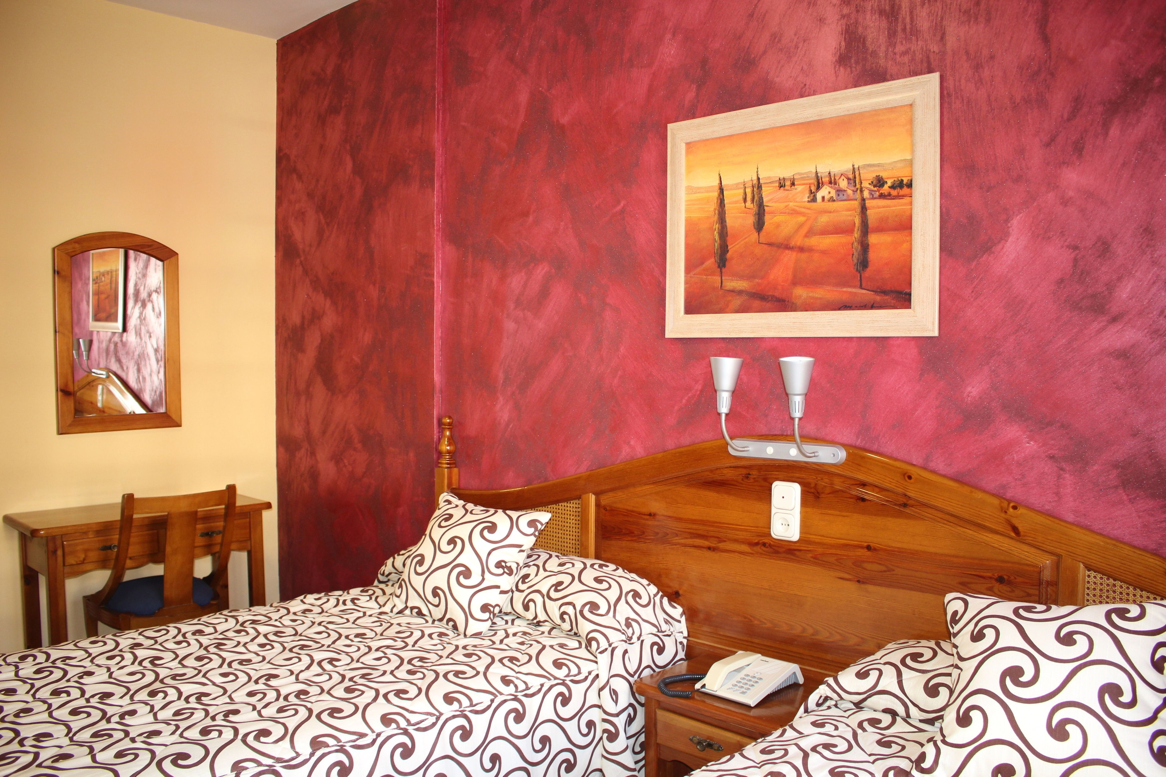 A bedroom with a wooden headboard, a bed with patterned bedding, a bedside table with a lamp, a telephone, and a painting on the wall.