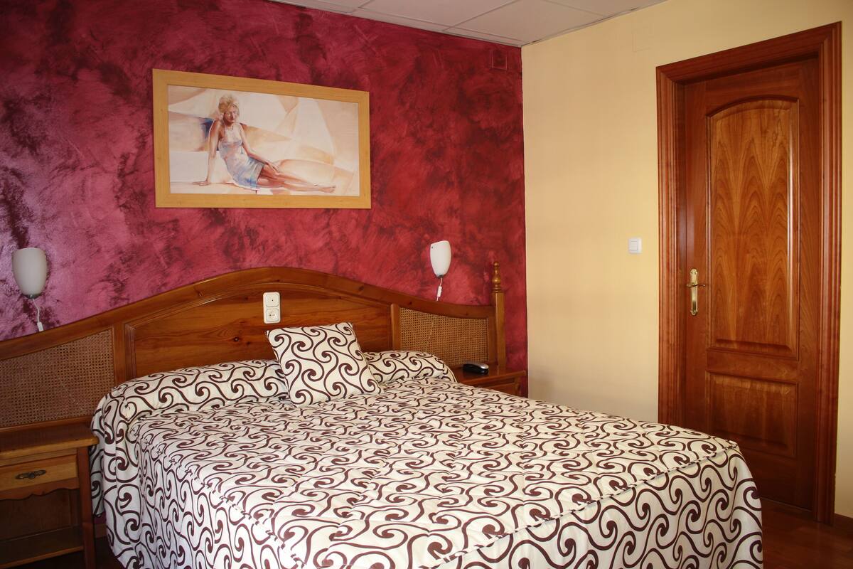 A bedroom with a wooden bed, a bedside table, a lamp, and a door.
