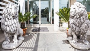Two stone lion statues in front of a building entrance with glass doors and potted plants. - JC Hotel (Rome)