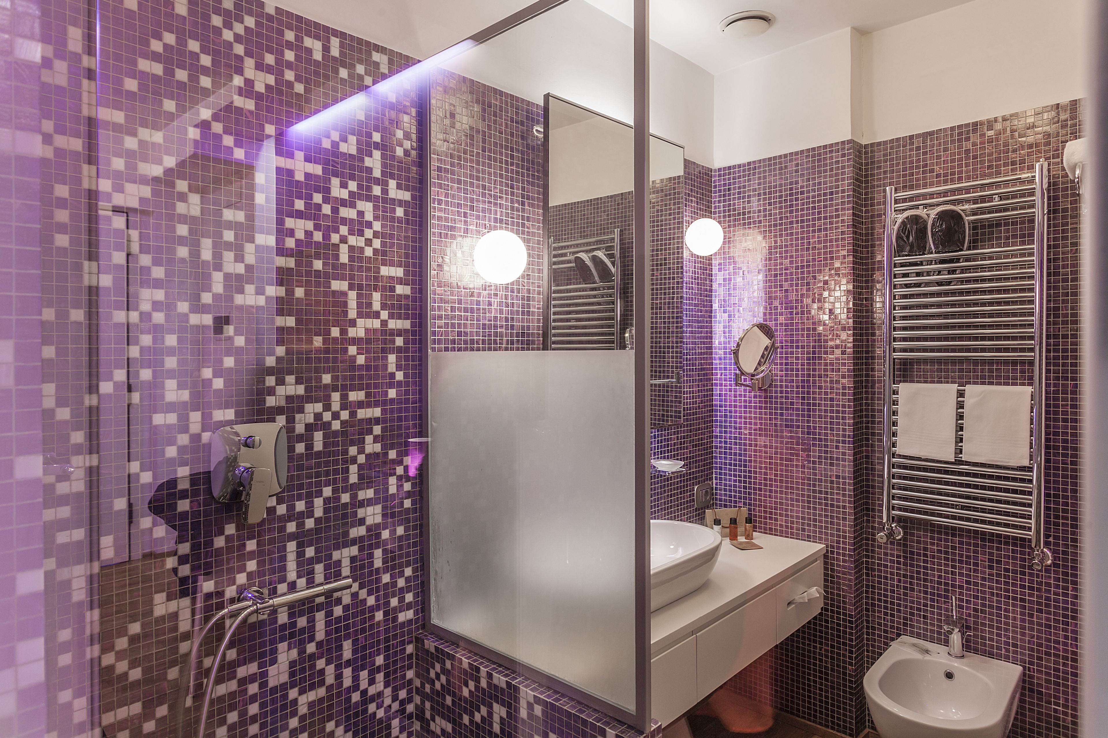 A bathroom with purple mosaic tiles, a glass shower enclosure, a sink, and a bidet.