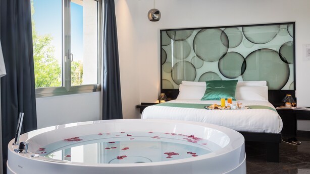 A modern hotel room with a large bathtub filled with water and rose petals, a neatly made bed with green and white bedding, and a large window with curtains. - JC Hotel (Rome)