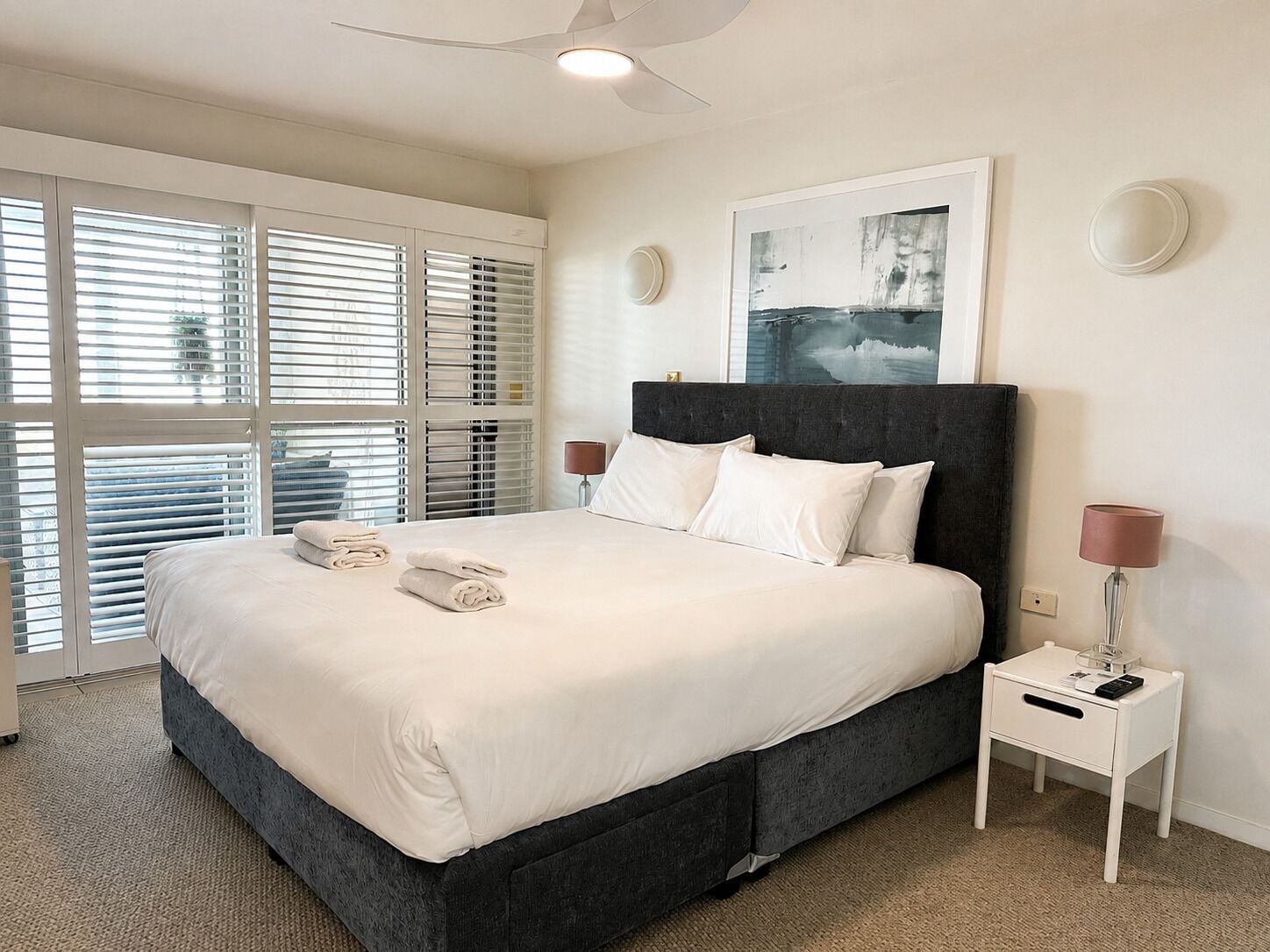 Ein Schlafzimmer mit einem Bett, zwei Nachttischen, einem Deckenventilator und einem großen Fenster mit Jalousien.