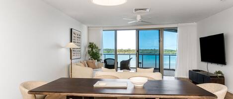 A modern living room with a large dining table, comfortable seating, a flat-screen TV, and a balcony with a view of the water.