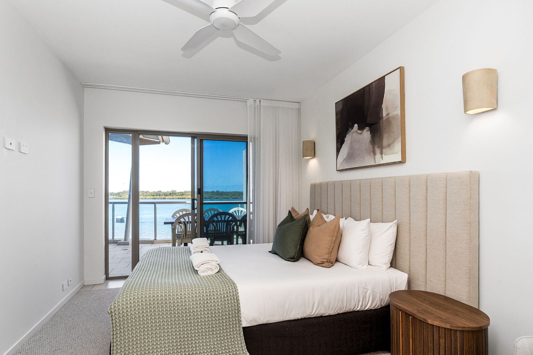 A hotel room with a bed, a ceiling fan, a painting, a lamp, and a view of the sea through a sliding door.
