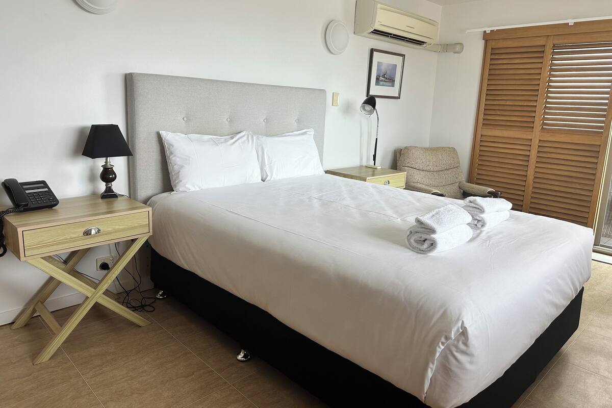 A neatly made bed with white linens, a bedside lamp, a telephone, and a small bedside table.