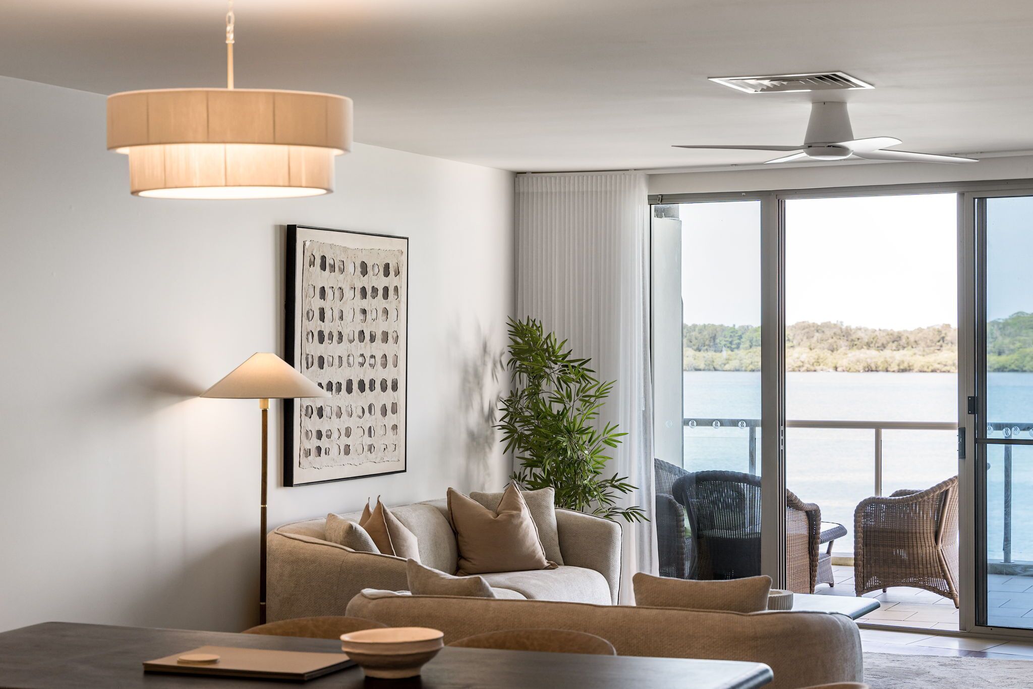 A modern living room with a sofa, a table, a lamp, and a large window with a view of the water.