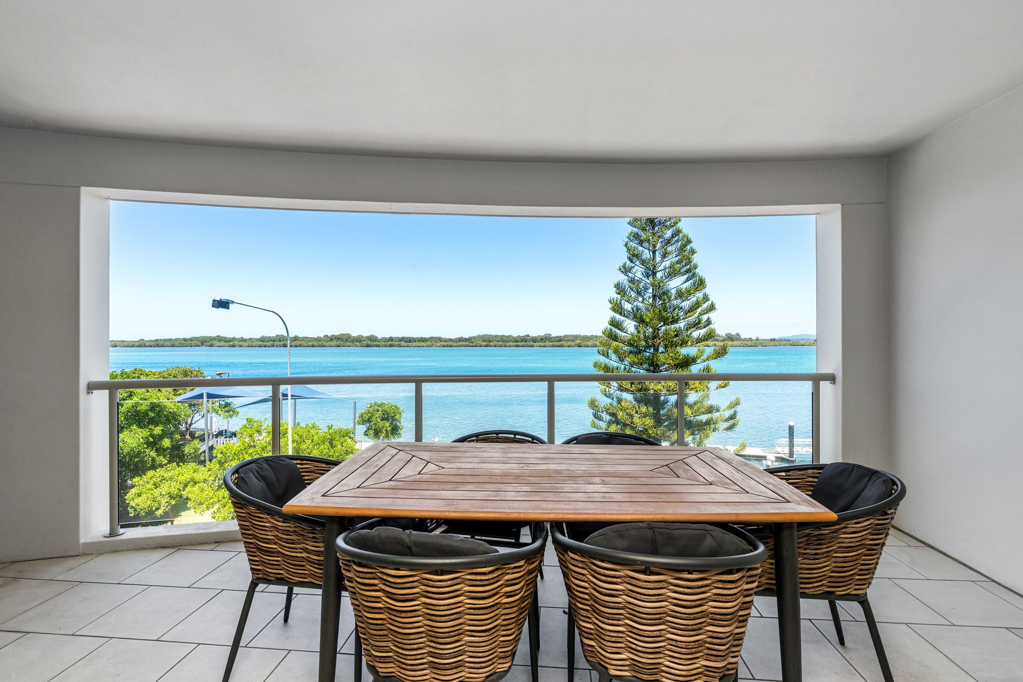 A balcony with a wooden table and wicker chairs, overlooking a body of water and greenery.