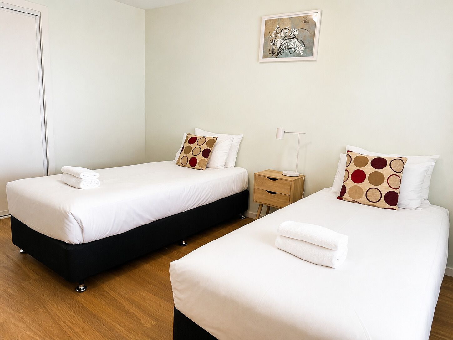 A hotel room with two beds, each with white bedding and patterned pillows, a bedside table with a lamp, and a framed artwork on the wall.