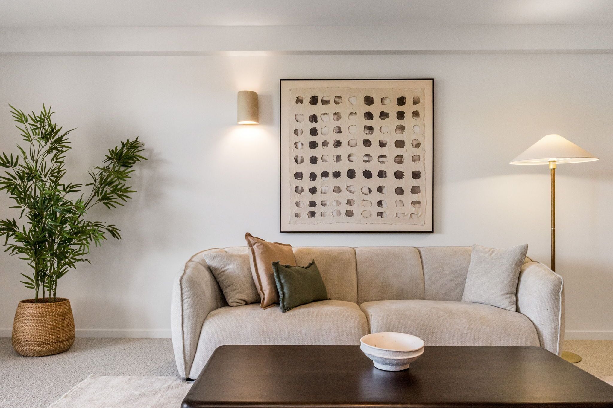 Un salon avec un canapé beige, une table basse en bois foncé, une plante en pot, un luminaire fixé au mur et une œuvre d’art abstraite encadrée.