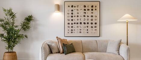 Un salon avec un canapé beige, une table basse en bois foncé, une plante en pot, un luminaire fixé au mur et une œuvre d’art abstraite encadrée.