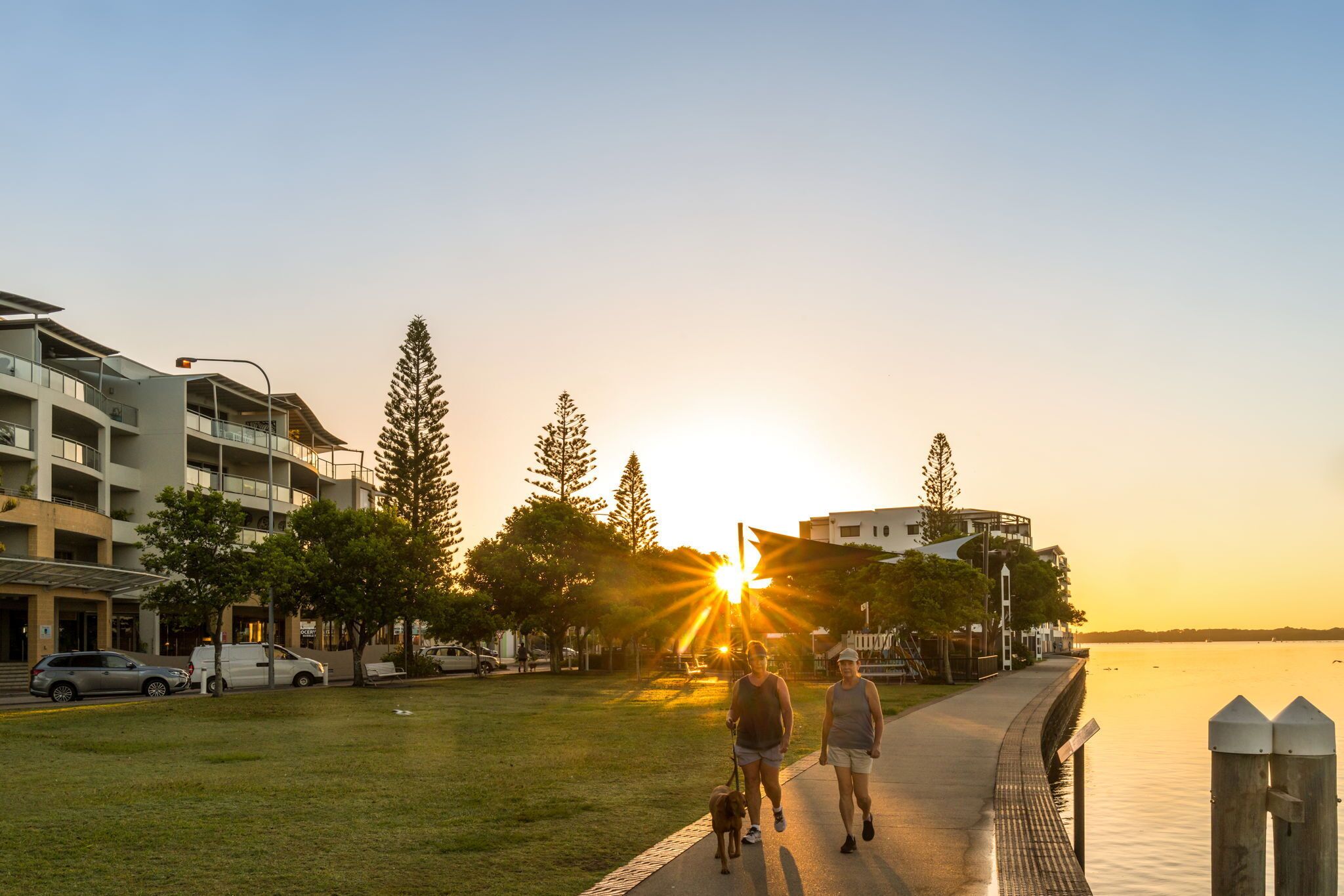 A waterfront promenade with a sunset, a pedestrian path, and modern buildings.