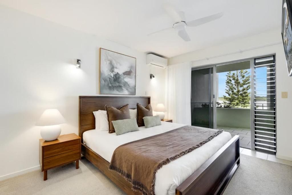 Une chambre avec un grand lit, un ventilateur de plafond, une table de chevet avec une lampe, un tableau au mur et une porte-fenêtre coulissante donnant sur un balcon.