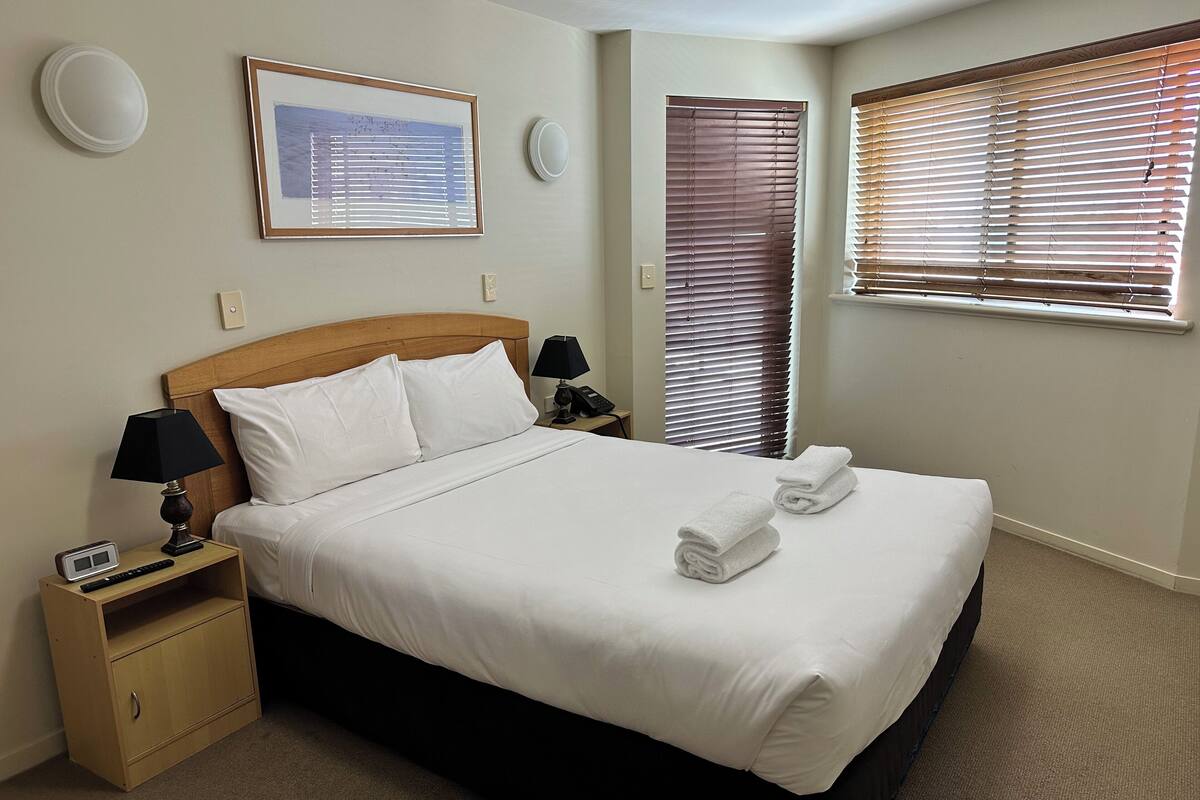 A neatly made bed with white linens, a wooden headboard, two bedside lamps, a framed picture on the wall, and a window with blinds.