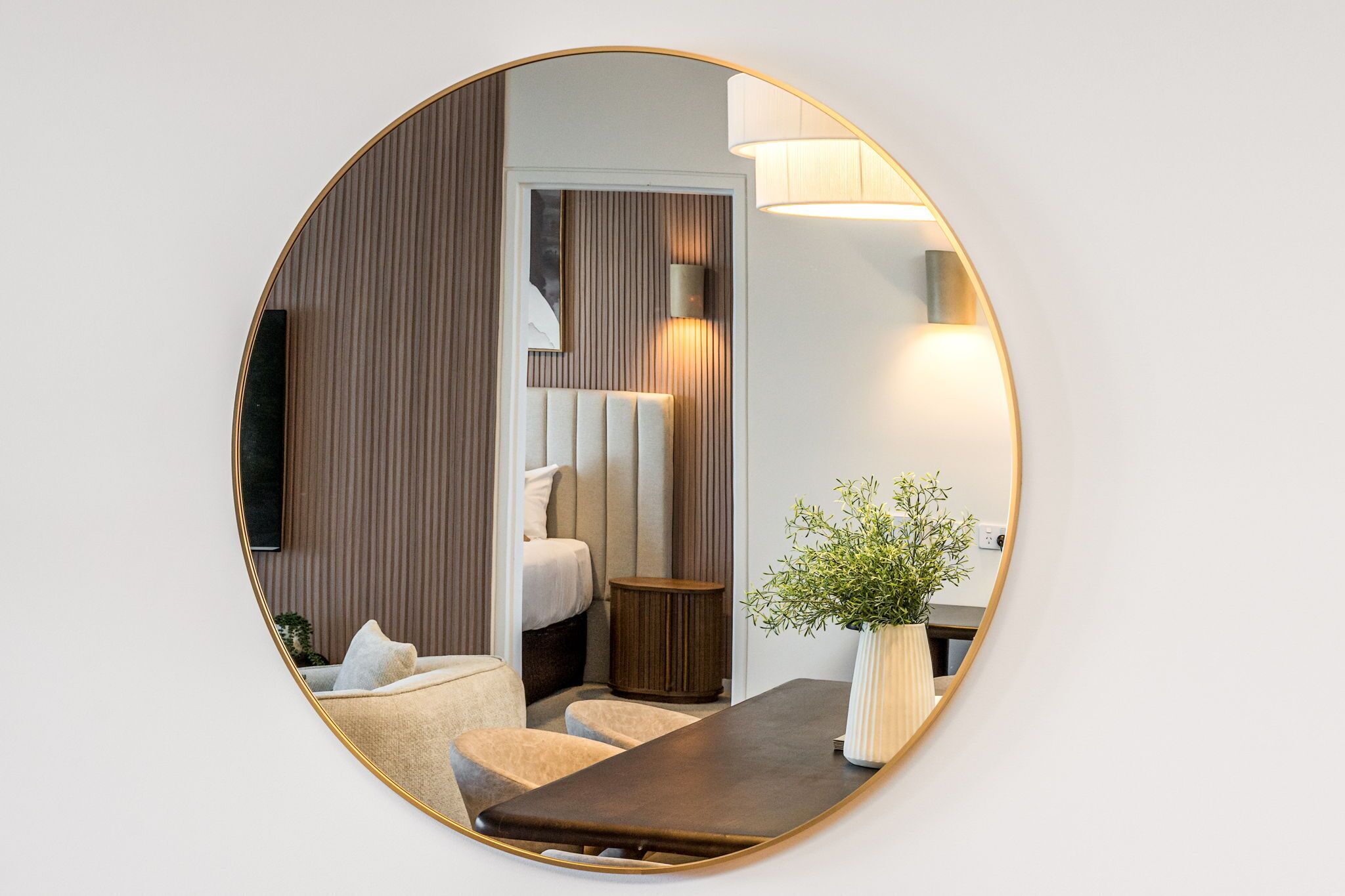 Een spiegel toont een moderne kamer met een bed, nachtkastje, lamp en een potplant.