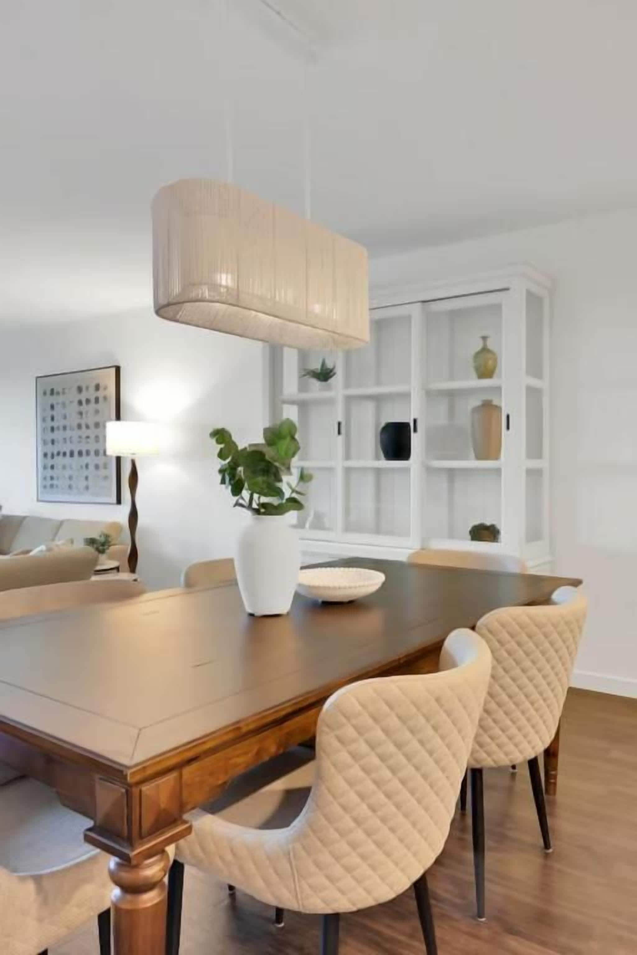 A dining area with a wooden table, two chairs, a hanging light fixture, a white bookshelf, and a potted plant.