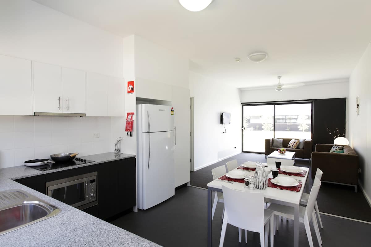 Una cocina moderna con armarios blancos, horno empotrado y refrigerador. Hay un comedor con mesa y sillas blancas, y un sofá en la sala de estar.