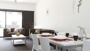 A modern dining area with a white table set for four, surrounded by white chairs. There is a sofa and a coffee table in the background. - Western Sydney University Village- Parramatta Campus (Parramatta)