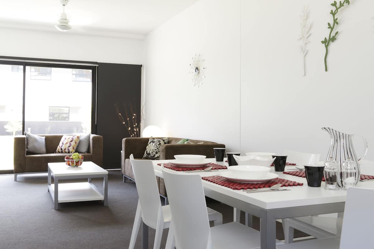 Un comedor moderno con una mesa blanca dispuesta para cuatro personas, rodeada de sillas blancas. Se aprecia un sofá y una mesa de centro al fondo.