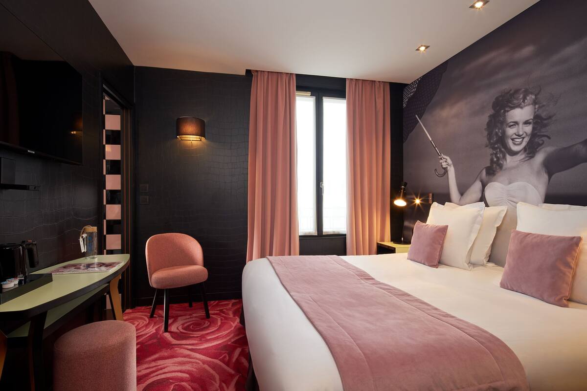 A hotel room with a large bed, a desk, a chair, a television, and a wall-mounted light.