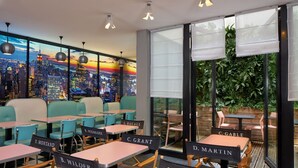 Un restaurant avec des tables et des chaises portant des noms, une fresque représentant un paysage urbain et de grandes fenêtres équipées de stores.