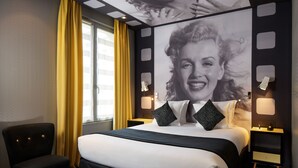 Une chambre d’hôtel avec un grand lit, une chaise, une table avec une tasse et une soucoupe, et une fenêtre avec des rideaux jaunes.