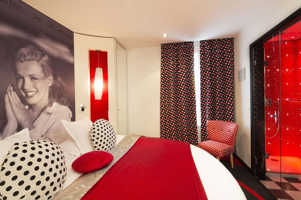 A modern hotel room with a bed, a polka-dotted chair, a red accent wall, and a patterned curtain.