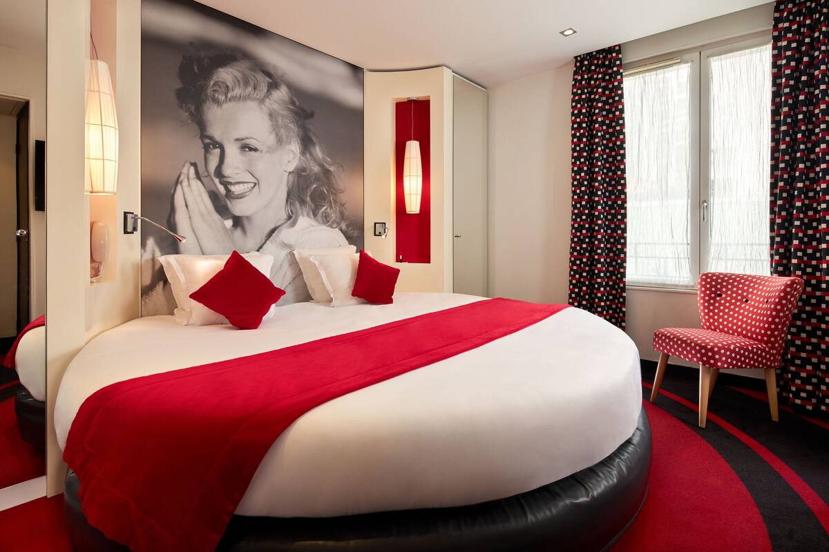 A large bed with red bedding and pillows, a red accent wall, a patterned chair, and a window with curtains.