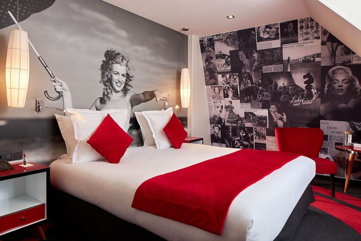 A hotel room with a large bed, red and white bedding, a red chair, and a wall decorated with black and white photos.